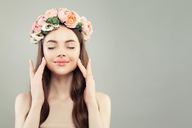 A woman with a headband of flowers and is gently massaging her cheeks while smiling. 