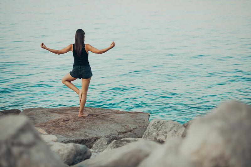 A woman standing on boulders near the sea and practing yoga.